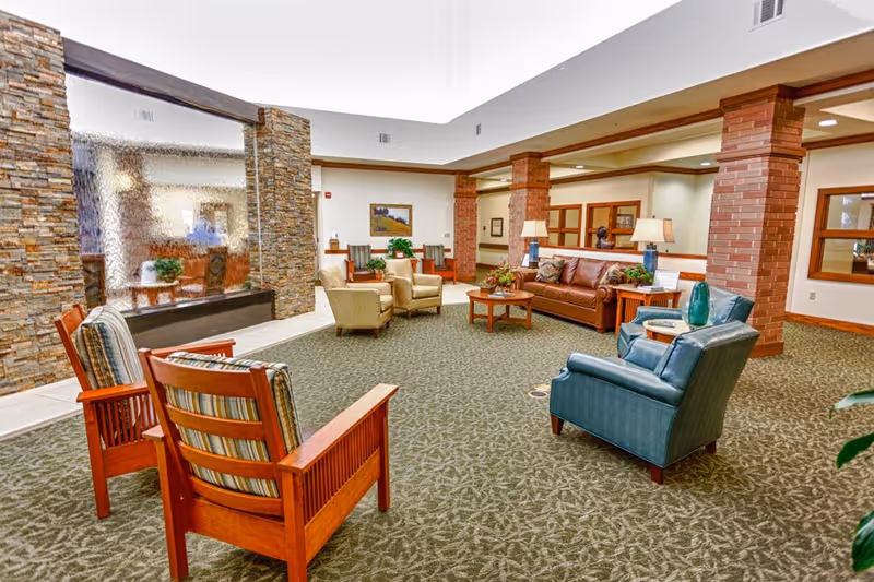 A spacious senior living facility common area with comfortable seating including wooden chairs with striped cushions, beige armchairs, a brown leather sofa, and a blue armchair. The room features a stone water wall, brick columns, carpeted floor, and warm lighting with table lamps and framed artwork on the walls.