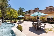 Outdoor courtyard with patio dining tables and umbrellas beside a decorative pool and landscaping in front of a Mediterranean-style building.