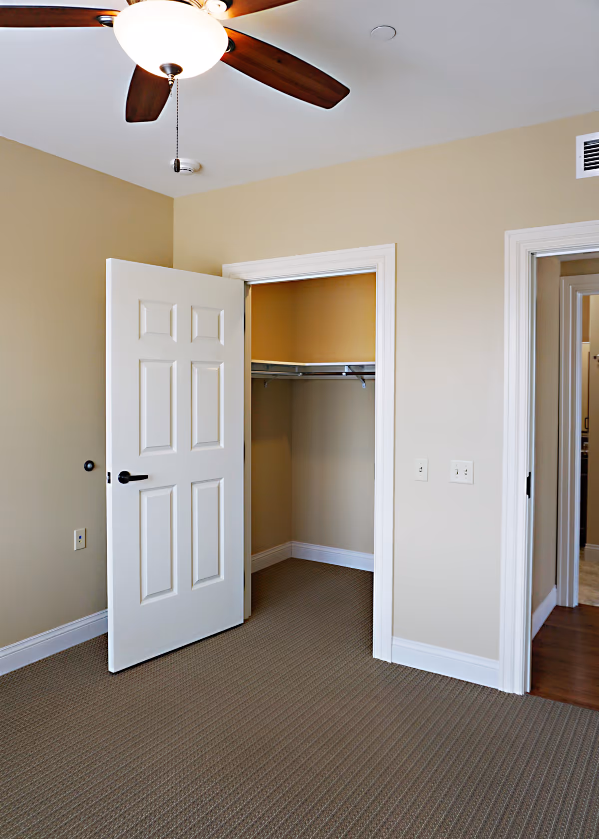 Empty bedroom with beige walls, a ceiling fan, an open closet, and a carpeted floor.