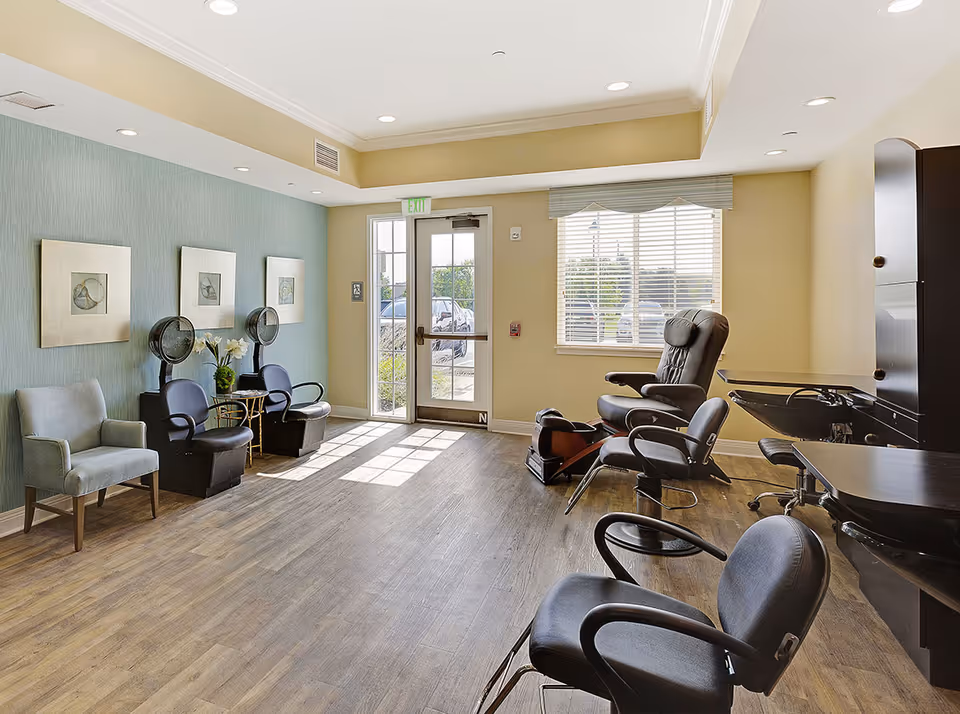 A bright salon area with styling chairs, hooded hair dryers, a pedicure chair, and seating by a glass door and window.