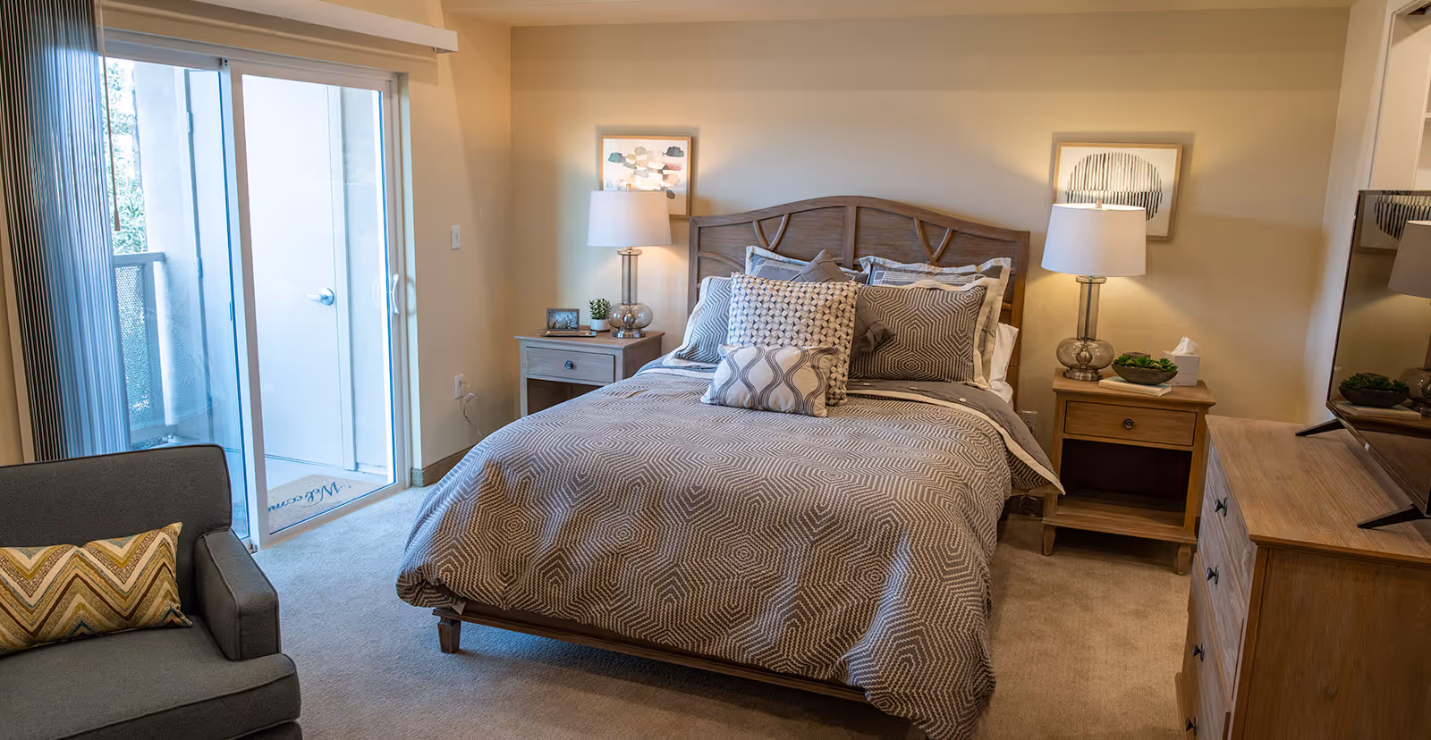 A cozy bedroom with a wooden bed frame and a neatly made bed featuring patterned bedding and multiple pillows. There are two wooden nightstands on either side of the bed, each with a lamp and decorative items. A sliding glass door leads to a balcony, and a gray armchair with a patterned pillow is positioned near the door. A wooden dresser with a TV on top is visible on the right side of the room.