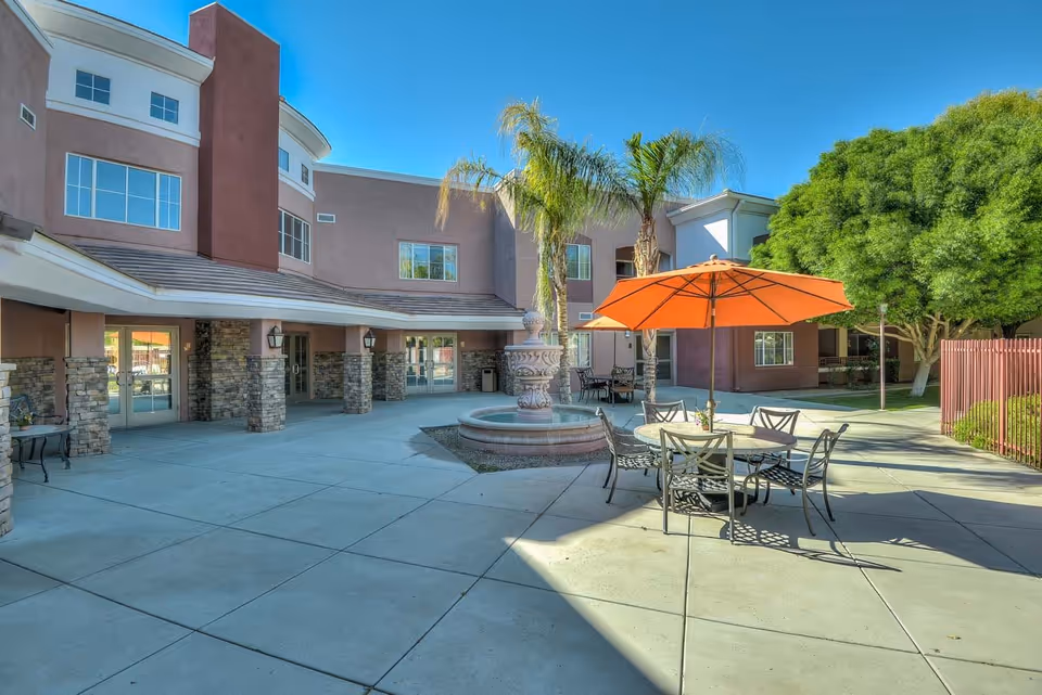 Outdoor courtyard with a central fountain, patio table and orange umbrella, palm trees, and a multi-story building facade.