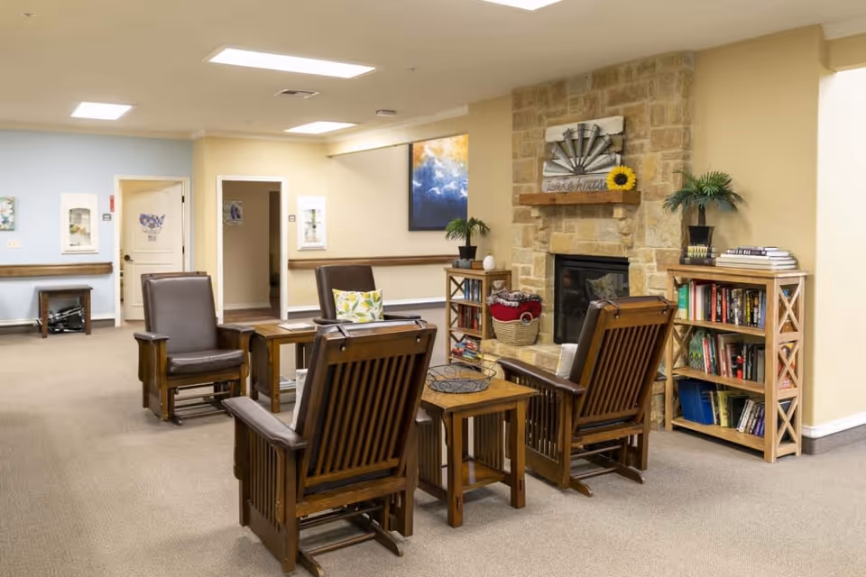 A cozy common area in an assisted living facility featuring four wooden armchairs with brown cushions arranged around a wooden coffee table. Behind the seating area is a stone fireplace with a decorative piece and a sunflower on the mantel. To the right of the fireplace are two wooden bookshelves filled with books and decorative items. The walls are painted in soft beige and light blue tones, and there is a large framed abstract painting on the wall. The room is well-lit with ceiling lights and has a carpeted floor.