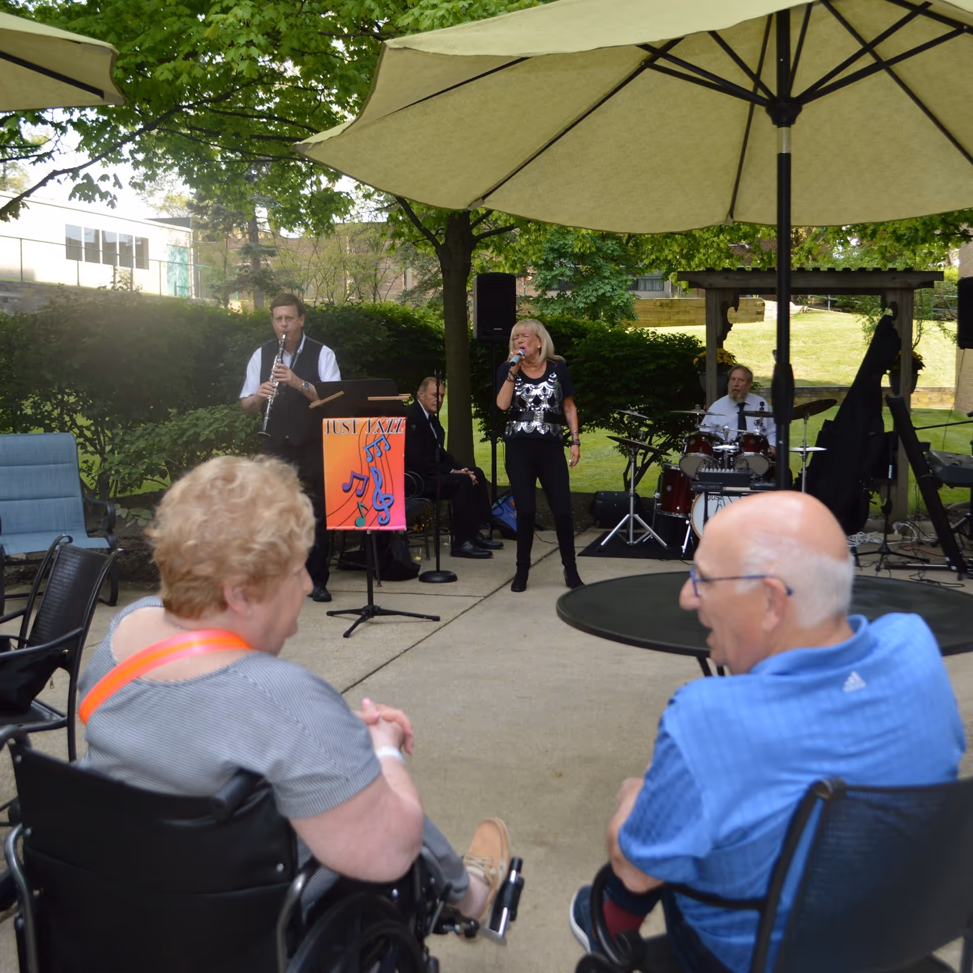 An outdoor patio area with two elderly people seated under large umbrellas watching a live jazz band perform. The band includes a clarinet player, a female singer, a drummer, and a pianist. The setting is surrounded by greenery and trees.