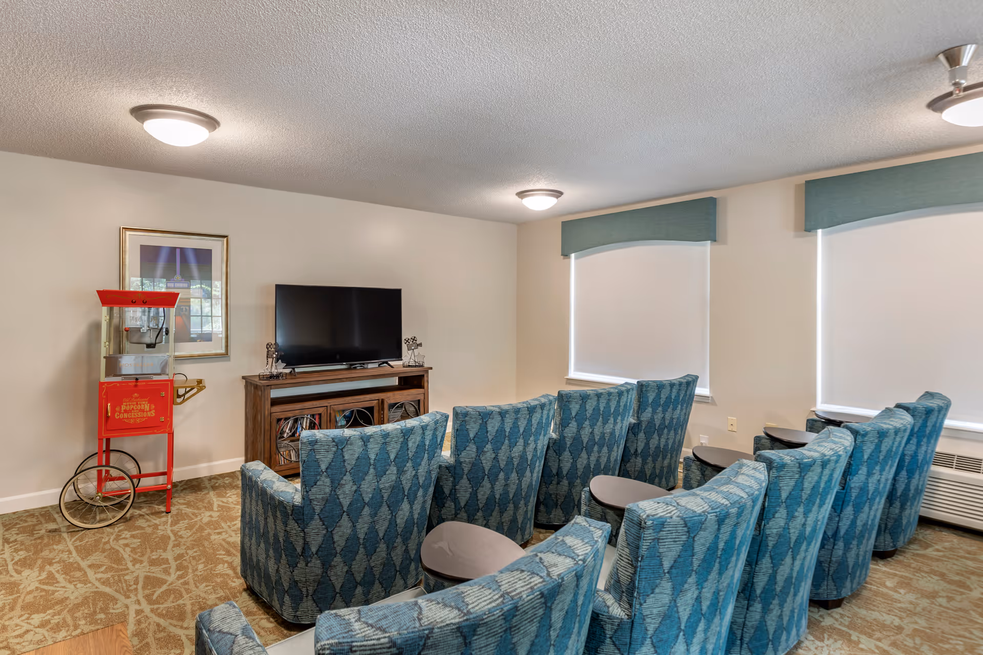 A small theater room with two rows of blue patterned armchairs facing a flat-screen TV on a wooden stand. There is a red popcorn machine on the left side of the room and two windows with white blinds and teal valances on the right wall. The room has beige walls and carpeted floor with a leafy pattern.