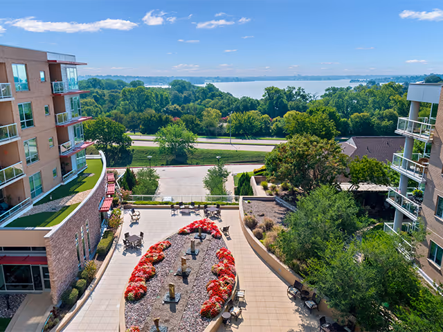 View from above of an outdoor courtyard area between two multi-story buildings with seating and decorative flower beds. Beyond the courtyard, there is a road, lush green trees, and a large body of water under a clear blue sky.