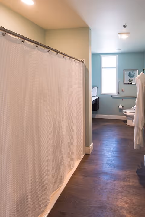 A bathroom with a white textured shower curtain on the left, a toilet and a white bathrobe hanging on the right, a window letting in natural light, a framed picture on the wall, and a wooden floor.