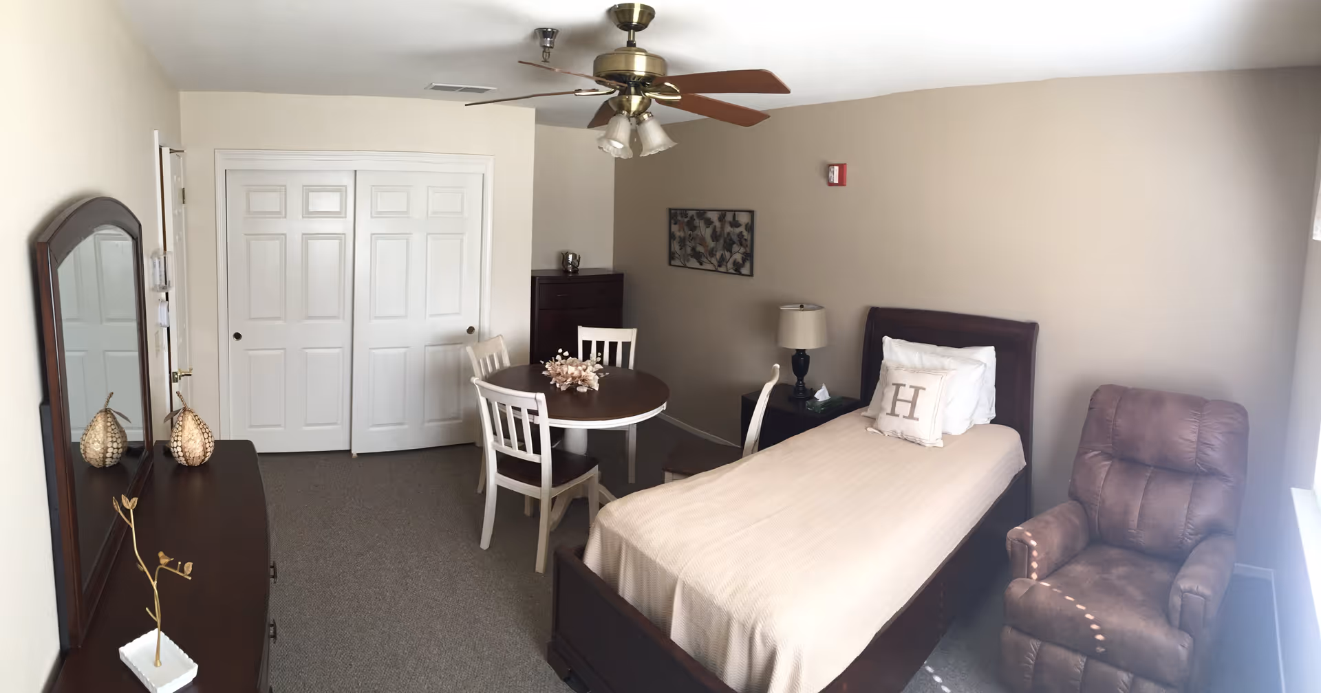 A senior living facility bedroom featuring a single bed with beige bedding and a pillow with the letter 'H'. The room includes a brown recliner chair near a window, a round wooden table with three chairs, a dresser with a mirror, and a ceiling fan with lights. The walls are light-colored, and there is a framed artwork on one wall.