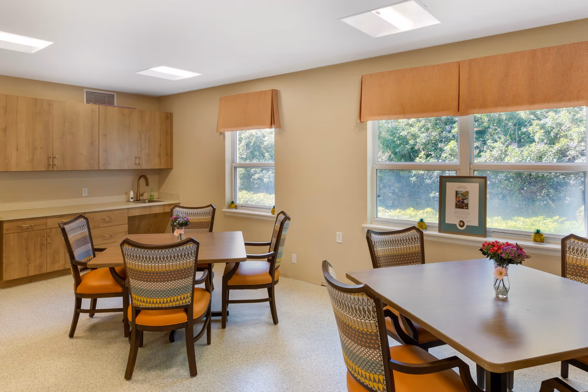 A bright room with two wooden tables, each surrounded by four chairs with patterned backs and orange seats. There are small flower arrangements on each table. The room has large windows with brown valances, letting in natural light and showing greenery outside. Along one wall are wooden cabinets and a countertop with a sink and soap dispenser.