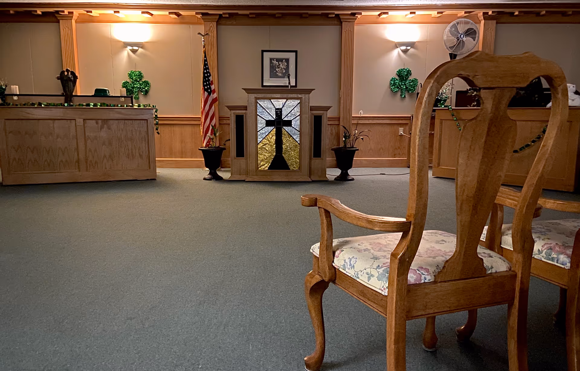 Wood-paneled interior meeting room with wooden chairs facing a central podium, an American flag, and shamrock decorations.