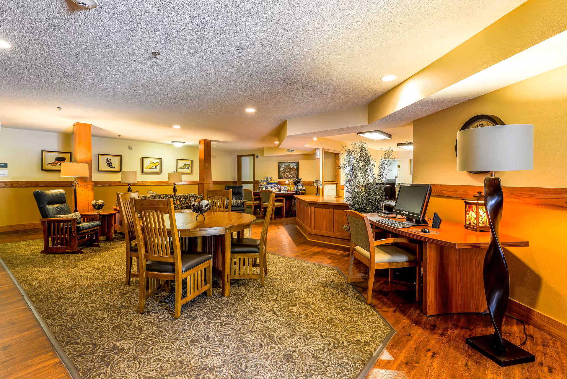 A cozy senior living common area featuring a round wooden table with four chairs, a computer desk with a chair and desktop computer, a tall floor lamp, and comfortable armchairs. The room has warm yellow walls, wooden accents, framed artwork on the walls, and a patterned carpet over hardwood flooring.