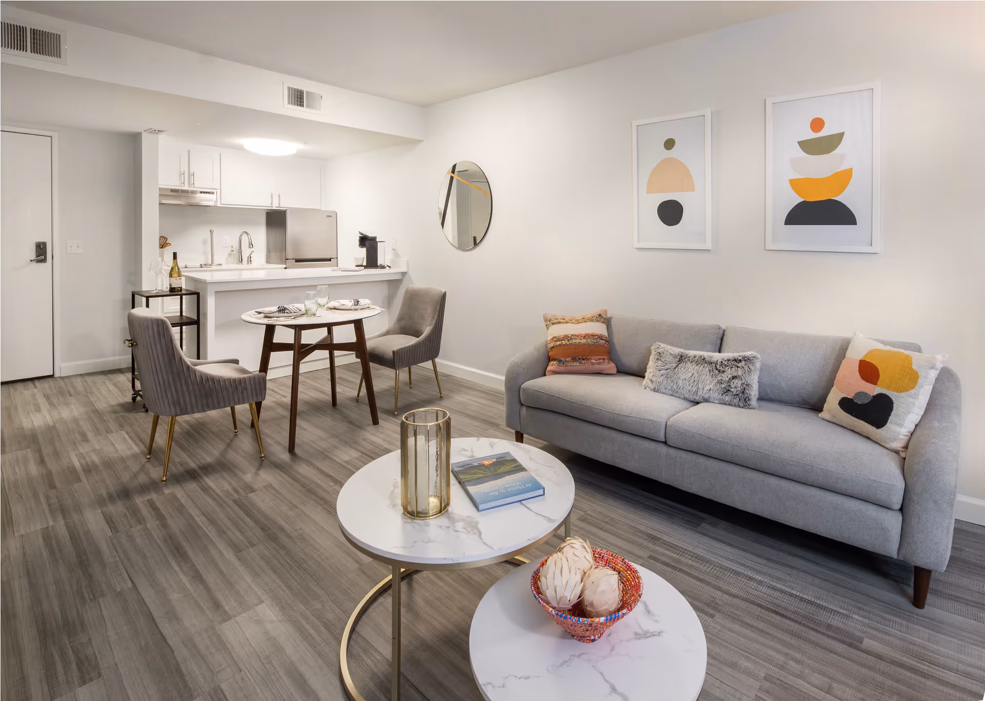 A modern living area with a gray sofa adorned with colorful pillows, two round white marble coffee tables with decorative items, a small round dining table set for two with gray upholstered chairs, and a compact kitchen with white cabinets and stainless steel appliances in the background. The walls are decorated with two abstract framed artworks and a round mirror.