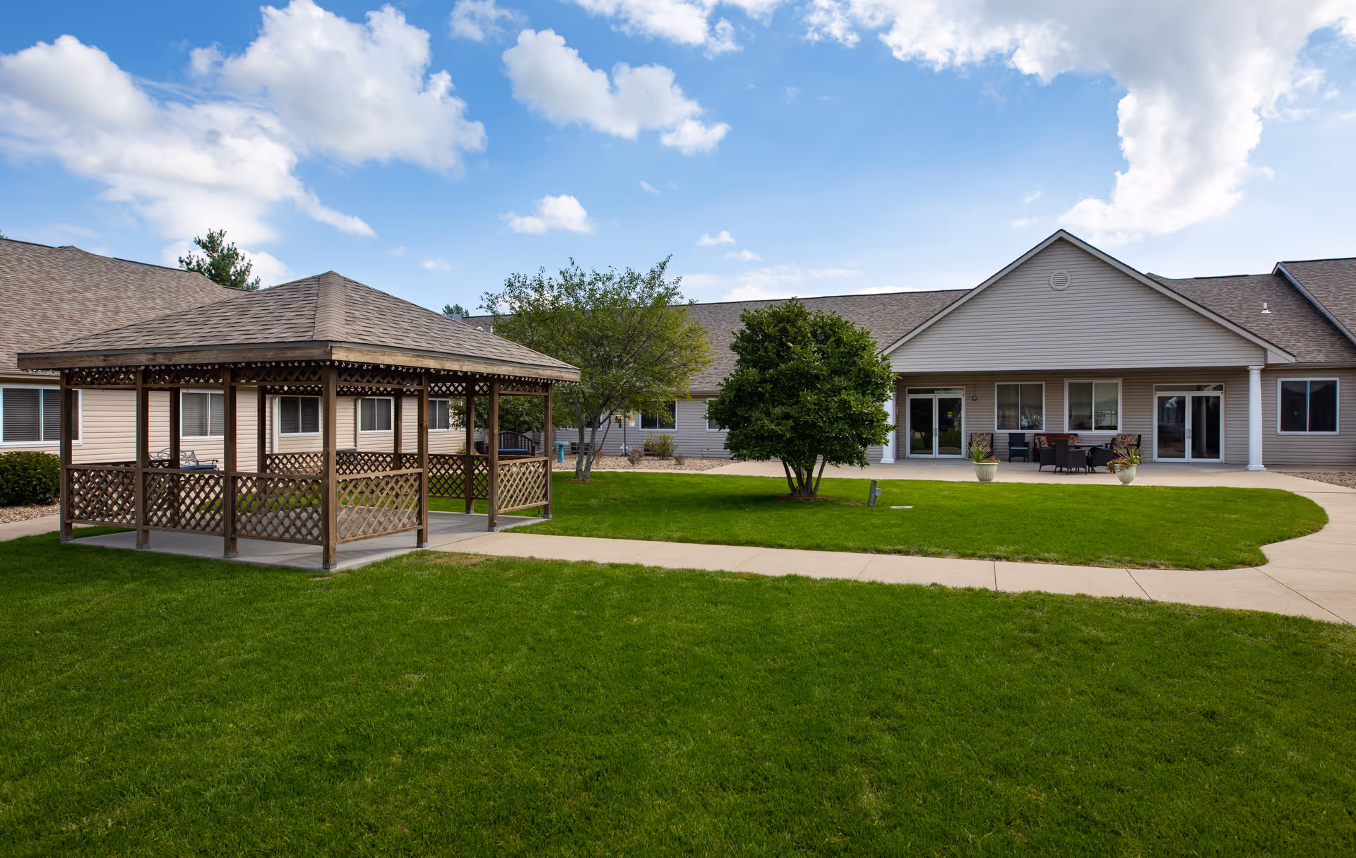 Outdoor view of a senior living facility with a wooden gazebo on a green lawn, a paved walkway, and a single-story building with beige siding and multiple windows and doors under a partly cloudy sky.