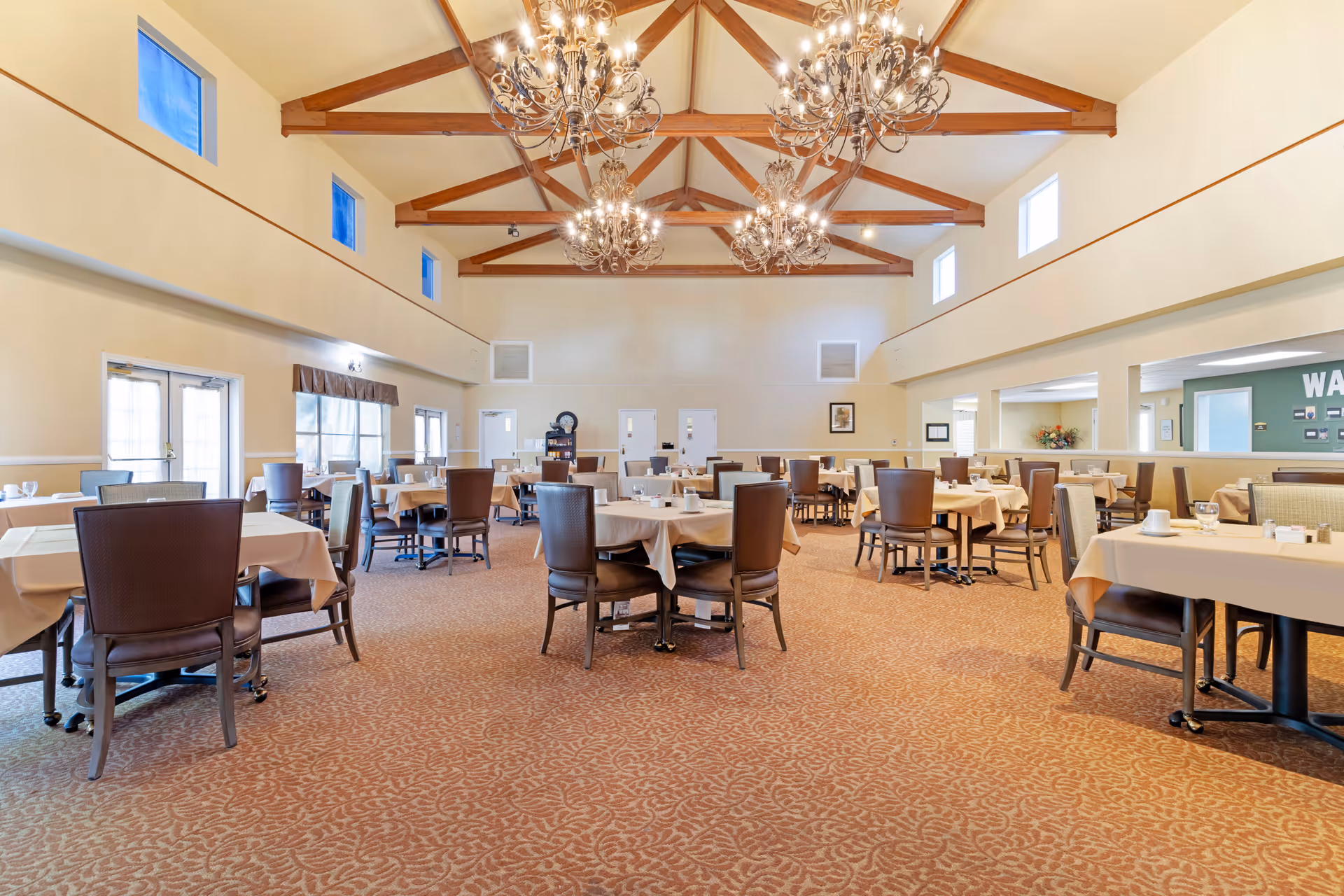 Spacious dining room with multiple tables covered in beige tablecloths and surrounded by brown chairs. The room features a high ceiling with exposed wooden beams and ornate chandeliers. There are windows near the ceiling and large windows on the side letting in natural light. The carpet has a patterned design in warm tones.