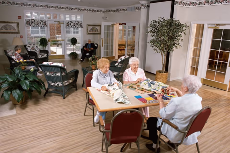 A bright common area in a senior living facility with three elderly women sitting around a table engaged in a quilting or sewing activity. In the background, two other elderly individuals are seated on cushioned wicker chairs near large windows and glass doors, with plants and framed artwork decorating the room.