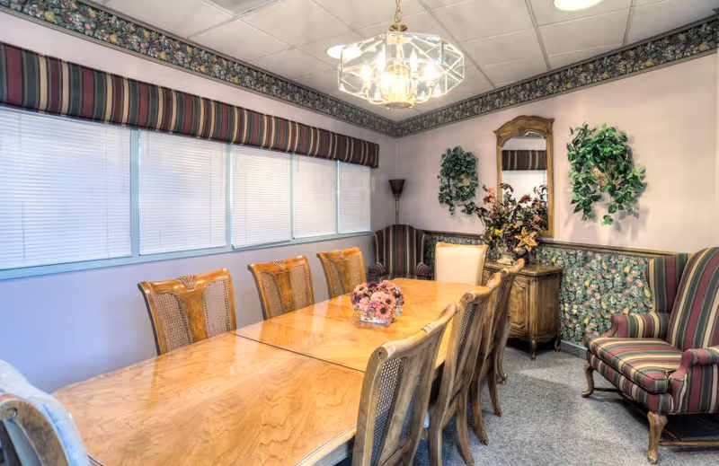 A dining room with a long wooden table surrounded by eight wooden chairs. The room has floral wallpaper borders and a striped valance over the windows with closed blinds. There is a chandelier hanging from the ceiling, a small wooden cabinet with a mirror above it, and two wall-mounted green plants. A striped armchair is positioned in the corner.