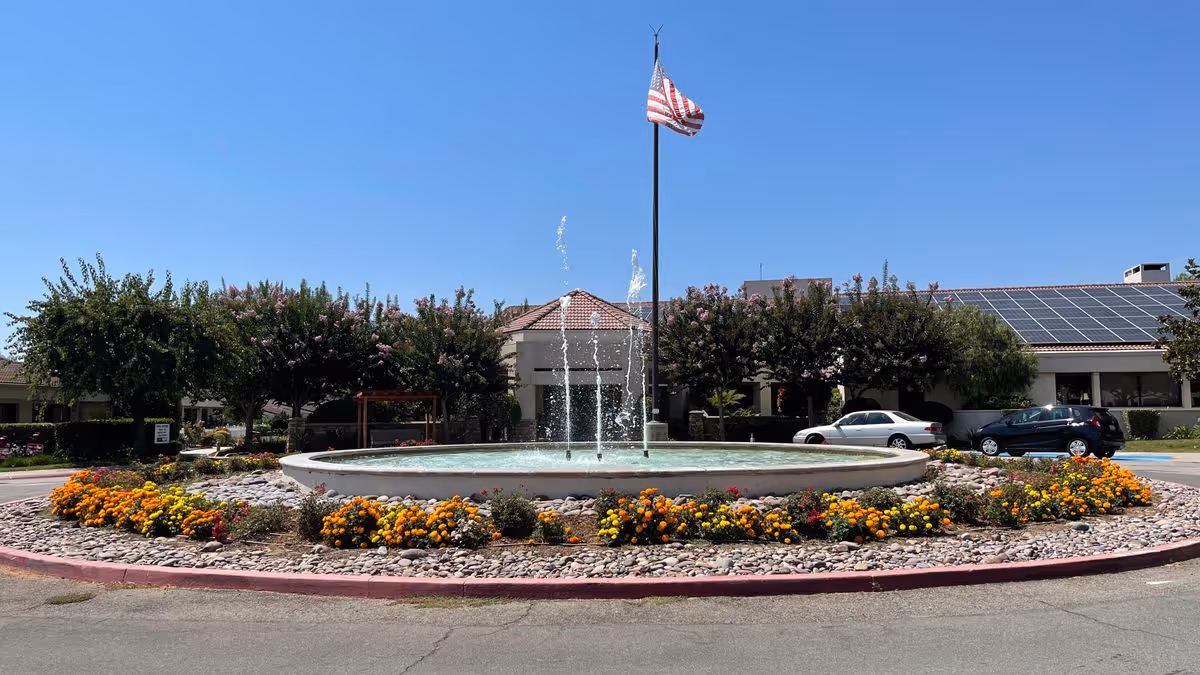 A circular fountain with jets and surrounding flowerbeds and an American flag in front of a low building with solar panels under a clear blue sky.