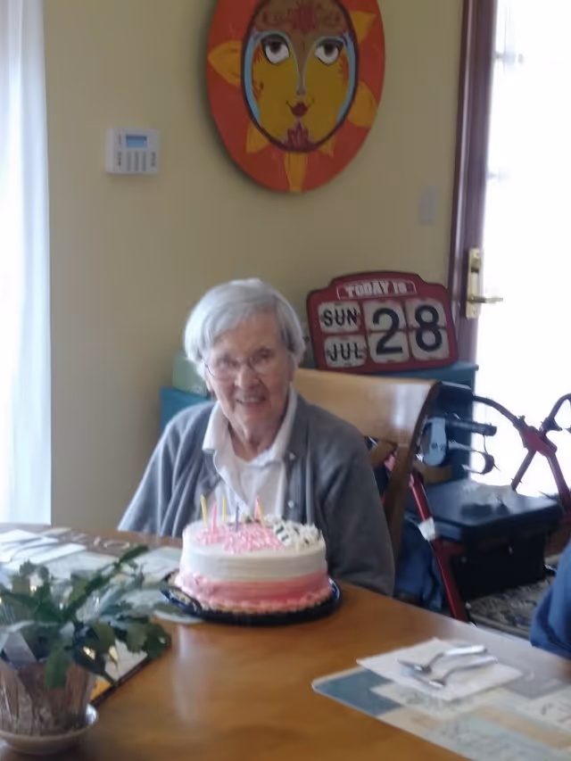 An elderly woman smiling at a table with a birthday cake in a dining area.