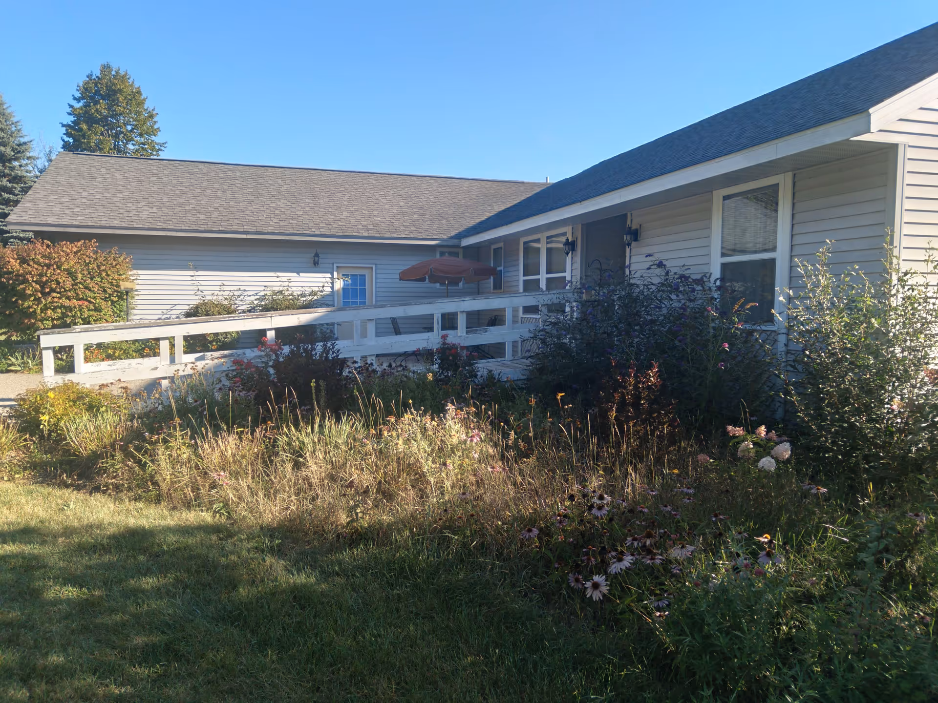 Exterior view of a single-story building with beige siding and a gray roof, featuring a white wooden ramp leading to the entrance. The surrounding area has a garden with various plants and flowers under a clear blue sky.