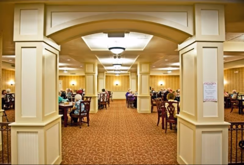 A spacious dining room in a retirement community with elderly residents seated at multiple wooden tables. The room features beige walls, carpeted flooring with a patterned design, and several large square columns with decorative molding. Ceiling lights provide warm illumination throughout the room.