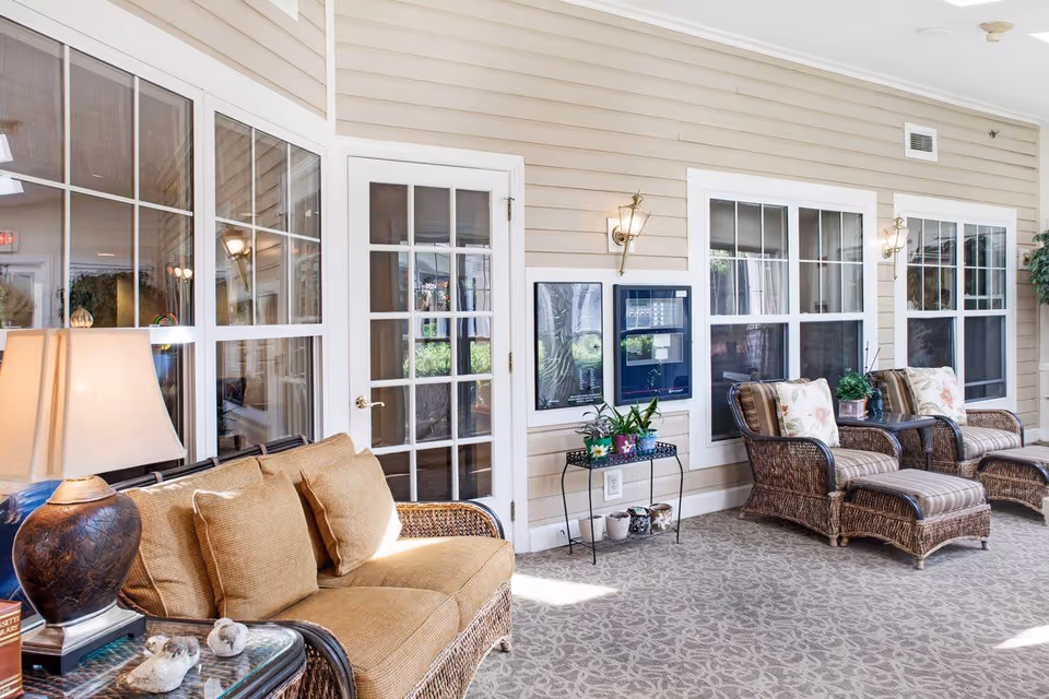 A bright and cozy indoor seating area with wicker furniture including a sofa and two armchairs with matching ottomans. The walls are beige with white trim around multiple windows and a glass-paneled door. There are decorative plants on a small black metal table and on the side tables. A table lamp with a brown base and beige shade is on a glass-top side table next to the sofa. The carpet has a subtle floral pattern, and wall-mounted light fixtures provide additional lighting.
