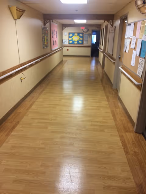 A long, empty hallway with wooden flooring and beige walls in a healthcare facility. The hallway has handrails on both sides, bulletin boards with various notices and papers, and ceiling lights. At the end of the hallway, there is a bulletin board with colorful decorations and an exit sign above a door.
