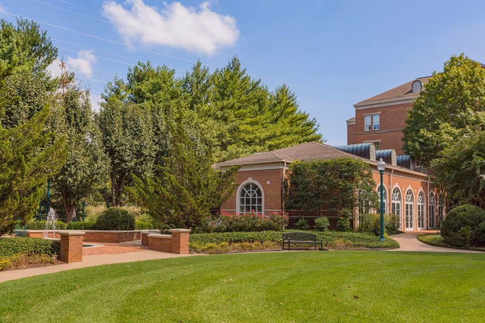 A well-maintained outdoor garden area with green grass, trees, and shrubs surrounding a brick building with large arched windows. There is a paved walkway, a bench, and a lamppost under a blue sky with some clouds.