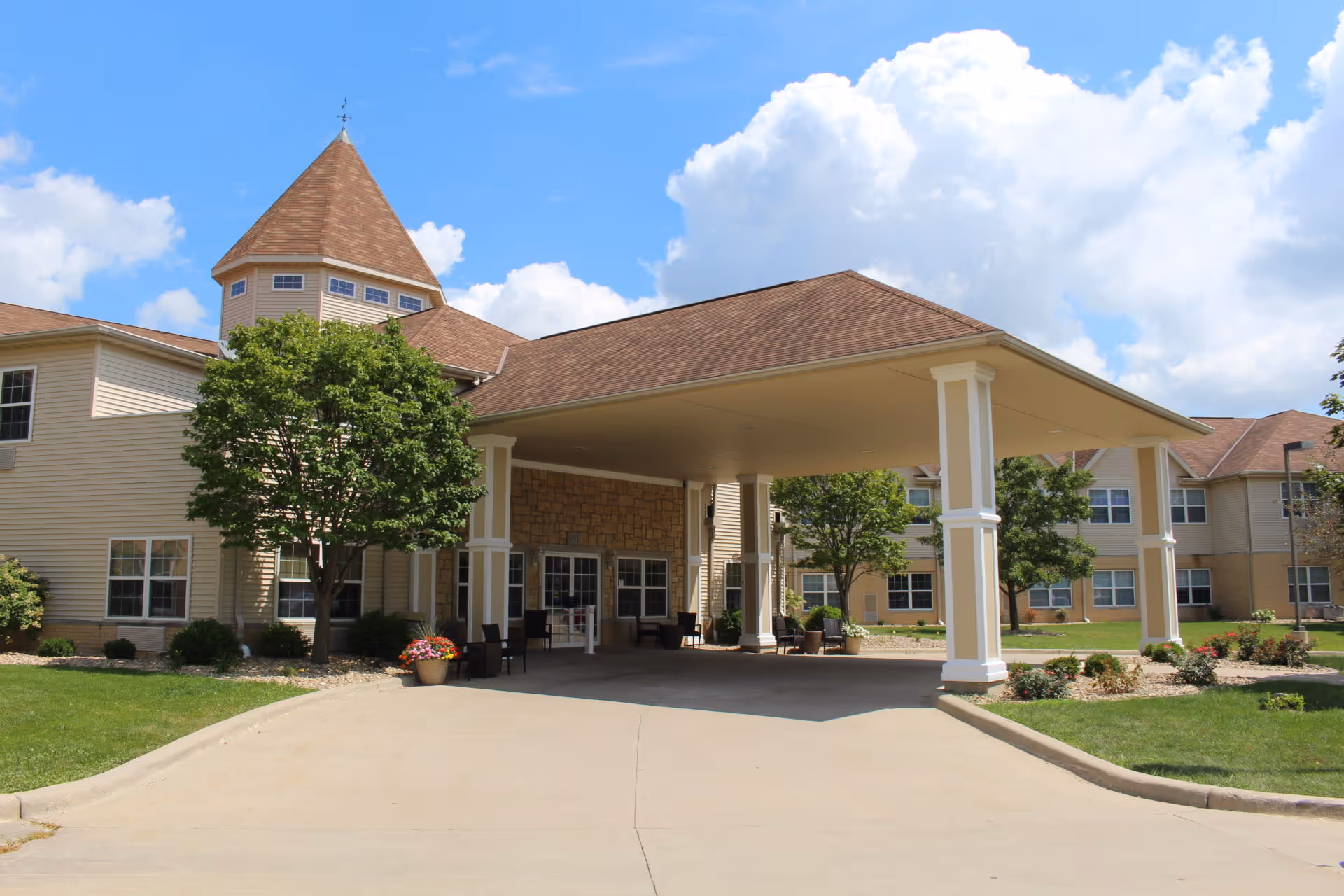 Exterior view of Evergreen Senior Living facility showing a large covered entrance with white pillars, beige siding, and a pointed turret roof. The building is surrounded by green lawns, trees, and flower beds under a partly cloudy blue sky.