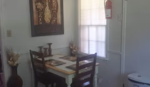 A small dining area with a wooden table set for two, featuring placemats and decorative vases. The room has a window with blinds, a fire extinguisher mounted on the wall, and a framed artwork hanging above the table.