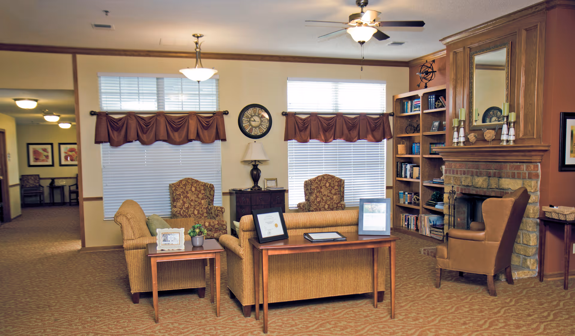 A cozy living room area in a senior living facility with two patterned armchairs, a sofa, a wooden side table with a small plant and picture frame, and a wooden console table with framed documents. The room features two large windows with brown valances, a ceiling fan with light, a brick fireplace with a large mirror above it, and a built-in wooden bookshelf filled with books and decorative items.
