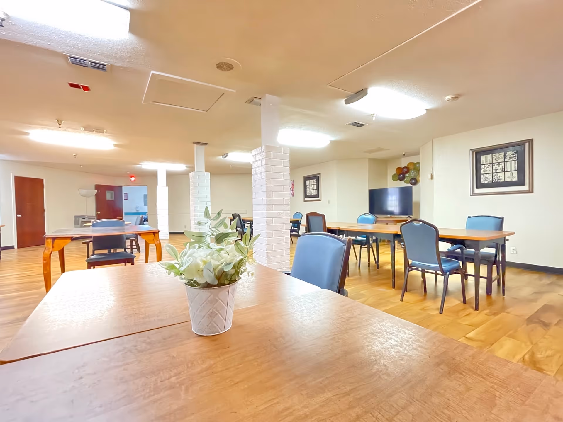 A well-lit common area with multiple wooden tables and blue cushioned chairs arranged around the room. A small white pot with green and white artificial flowers is placed on the nearest table. The room has white walls, framed artwork, and a flat-screen TV mounted on a stand. The floor is wooden, and there are white brick pillars in the center of the room.