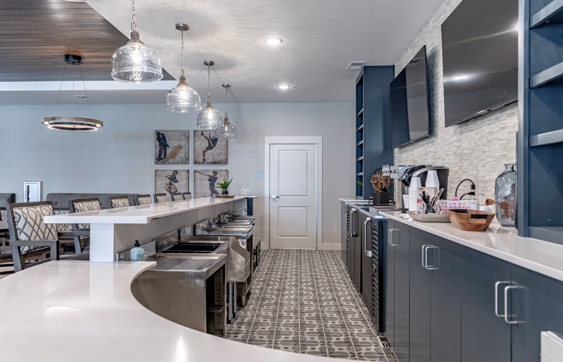 Modern kitchen area with a long white countertop, blue cabinets, patterned tile floor, and three hanging glass pendant lights. There are two flat-screen TVs mounted on the wall above the counter, and several chairs with patterned upholstery are visible in the background. The space is clean and well-lit with recessed ceiling lights.