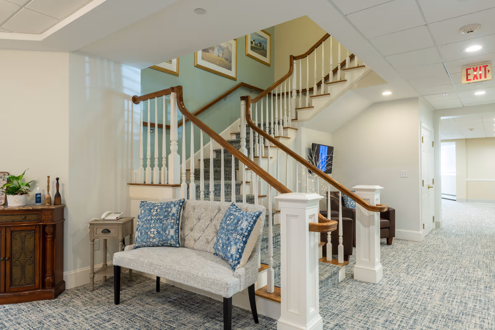 Interior view of a senior living facility hallway featuring a carpeted staircase with wooden handrails and white balusters. A cushioned bench with two blue patterned pillows is placed near the staircase. There is a small side table with a telephone and a decorative cabinet with plants and decorative items. The hallway is well-lit with ceiling lights and has light-colored walls and carpeted floors. An exit sign is visible in the background.