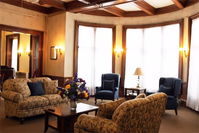Sunlit common sitting room with patterned sofas, two blue armchairs, a coffee table with a floral arrangement, and large bay windows.