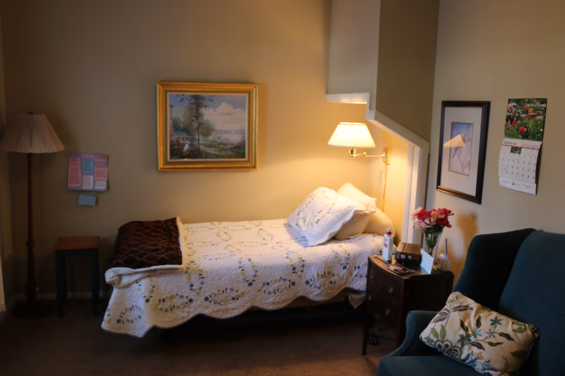 A cozy bedroom with a single bed covered in a white quilt with floral patterns. A bedside table next to the bed holds a lamp, flowers, and some personal items. There is a green armchair with a floral cushion, a floor lamp, a small side table, framed artwork on the walls, and a calendar.
