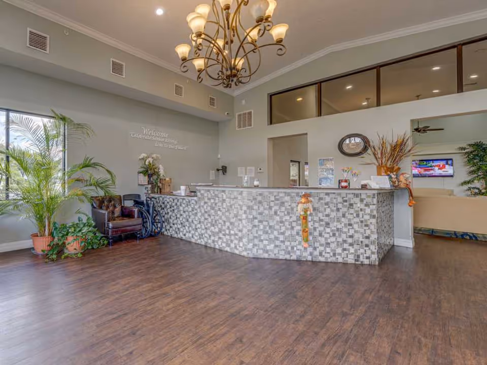 Reception area of Regency Park Assisted Living & Memory Care featuring a tiled front desk with decorative fall-themed scarecrow figures, a chandelier overhead, a leather chair and wheelchair near a window with potted plants, and a seating area with a television in the background.
