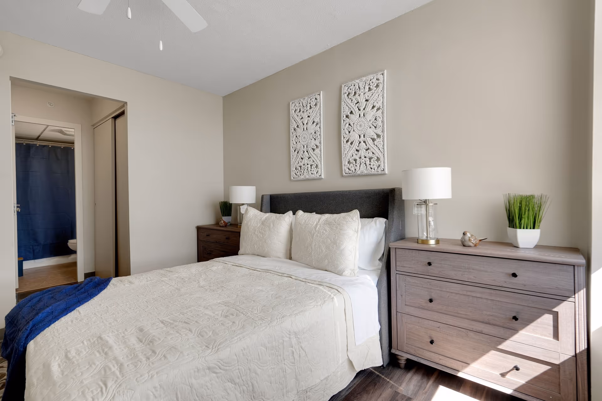 A neatly made bed with white bedding and three pillows in a bedroom. On either side of the bed are wooden nightstands with white lamps and decorative items. Two ornate white wall hangings are above the bed. A doorway leads to a bathroom with a blue shower curtain visible. Sunlight streams into the room from the right side.