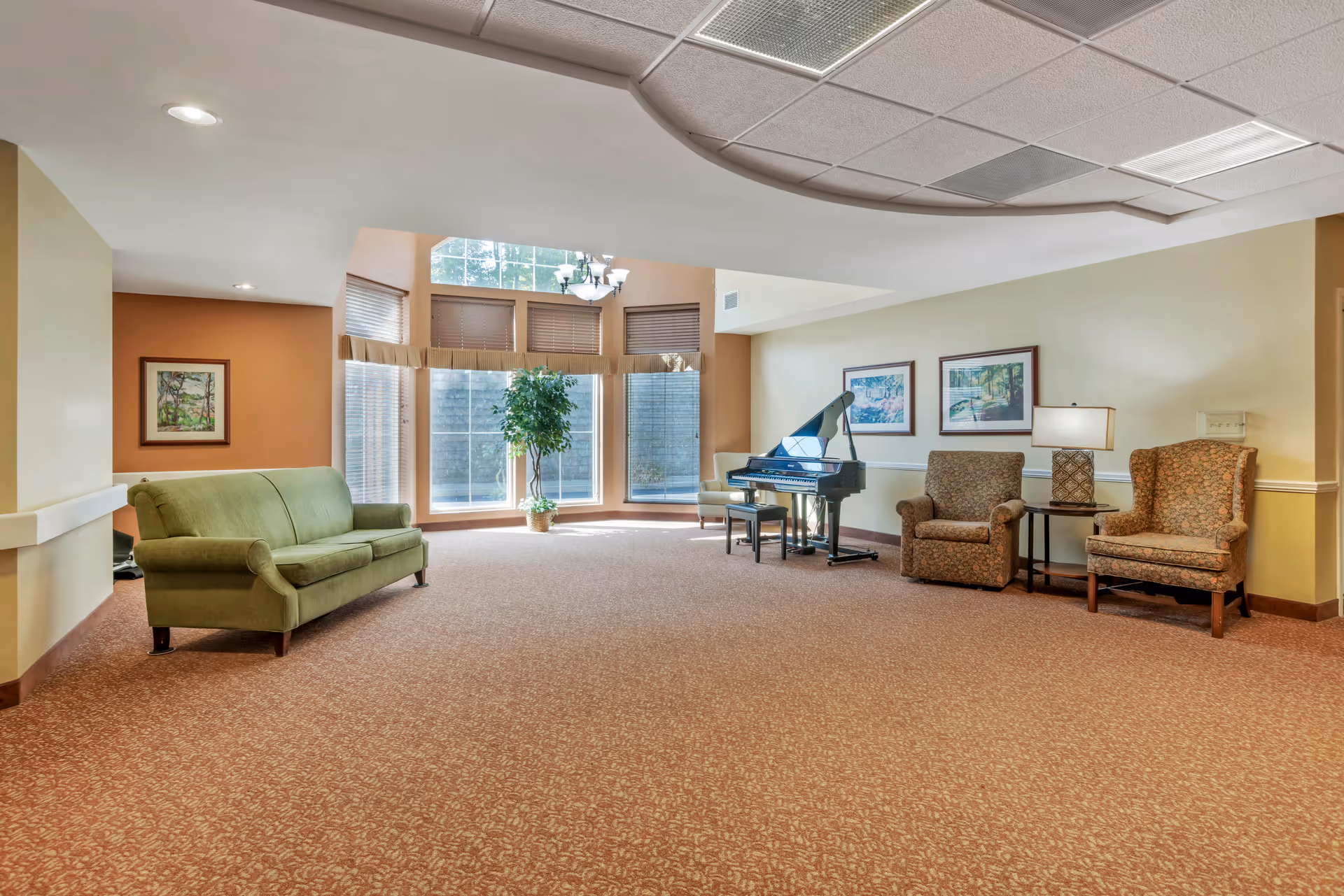 A spacious living room area with a green sofa on the left, two patterned armchairs on the right separated by a small table with a lamp, and a black grand piano with a bench near large windows with blinds. The room has beige walls, carpeted floor, and framed artwork on the walls.