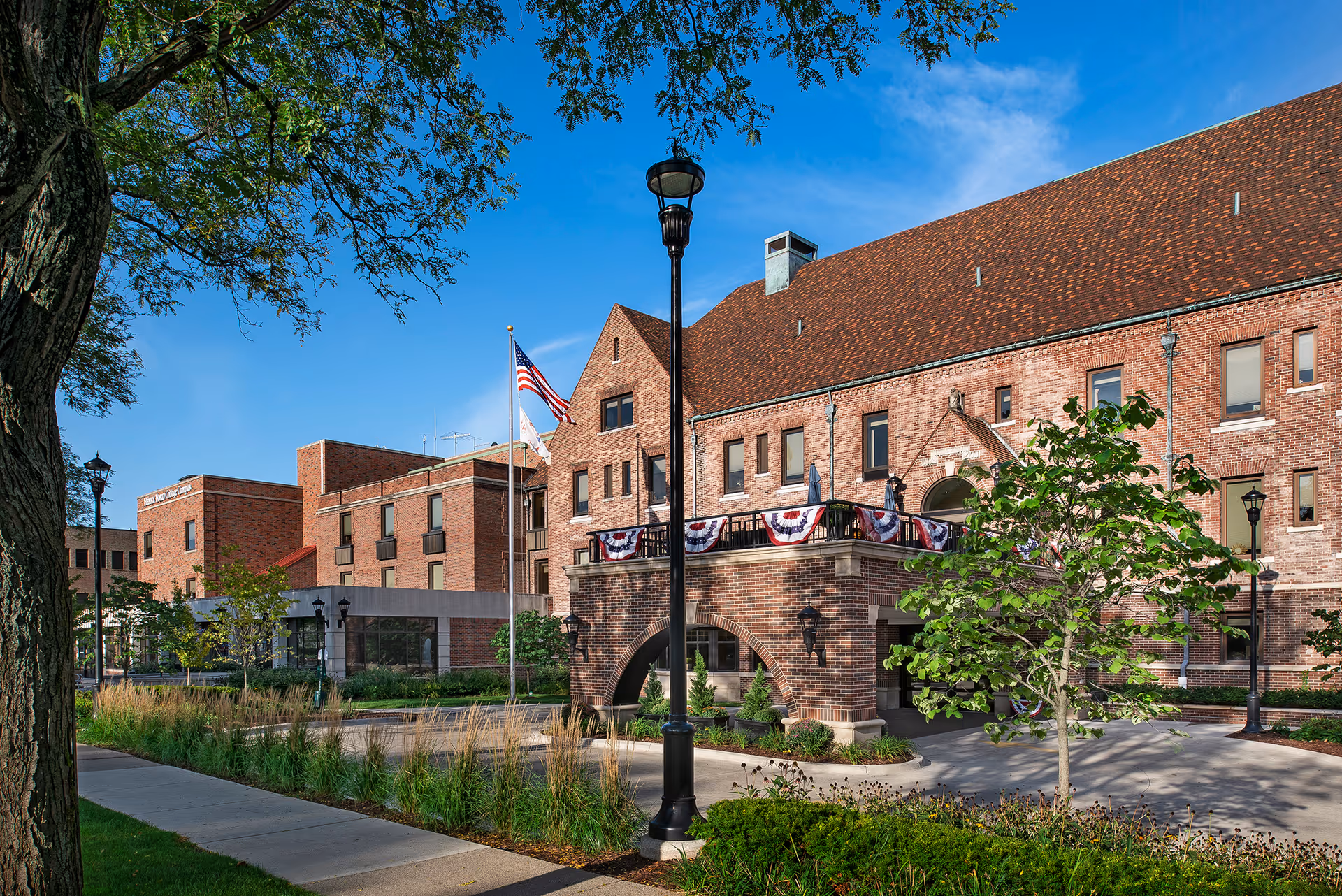 Exterior view of a large brick building with multiple windows and a steep roof, decorated with patriotic bunting. There are flagpoles with the American flag and another flag, a sidewalk, street lamps, trees, and landscaped greenery under a clear blue sky.