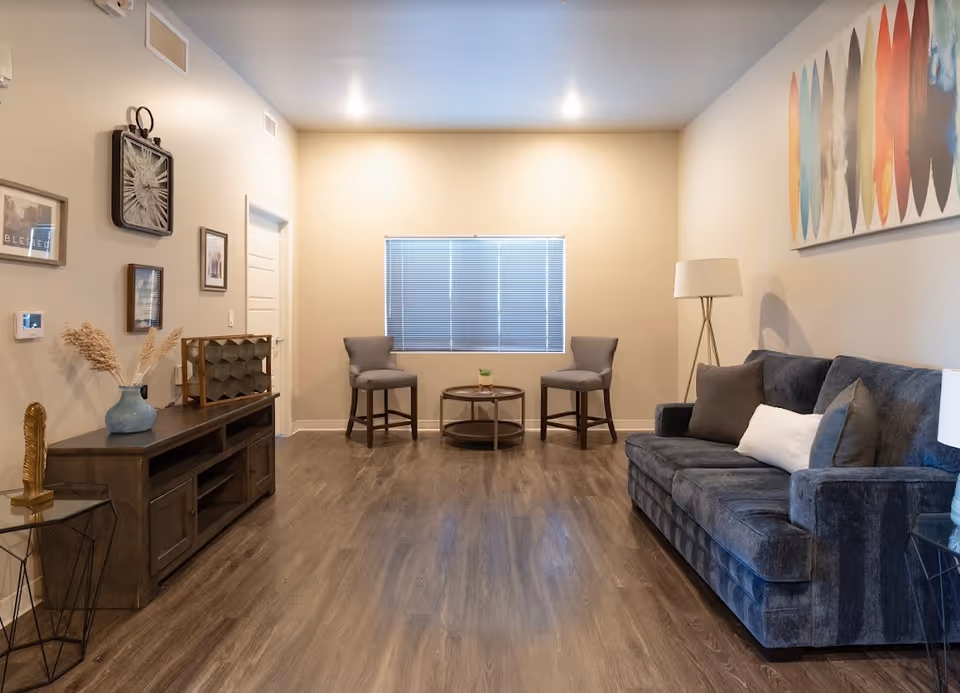 A cozy living room with a dark gray sofa adorned with pillows on the right, two gray chairs and a round coffee table in front of a window with closed blinds. The room has wooden flooring, a floor lamp, a side table with a blue vase holding dried plants, a wooden console table with decorative items, and colorful abstract artwork on the wall.
