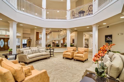 Spacious and well-lit senior living facility common area with beige and tan sofas and armchairs arranged around a central carpeted floor. The room features white columns, a high ceiling with a second-floor balcony railing, and decorative plants including orange flowers on a side table. A staircase and chandelier are visible in the background.