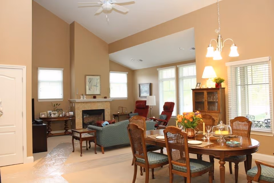 A cozy living and dining area with beige walls and carpet. The dining table is set with plates, glasses, and a vase of orange tulips. The living area features a green sofa, two red armchairs, a fireplace with a mantel decorated with framed photos and candles, and large windows letting in natural light. A wooden cabinet with glass doors and a lamp is also visible.