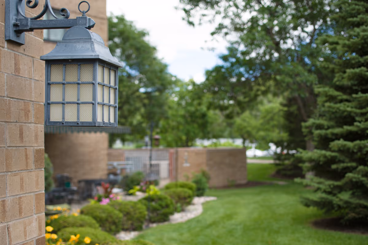 A metal exterior lantern mounted on a brick wall beside a landscaped lawn and trees.