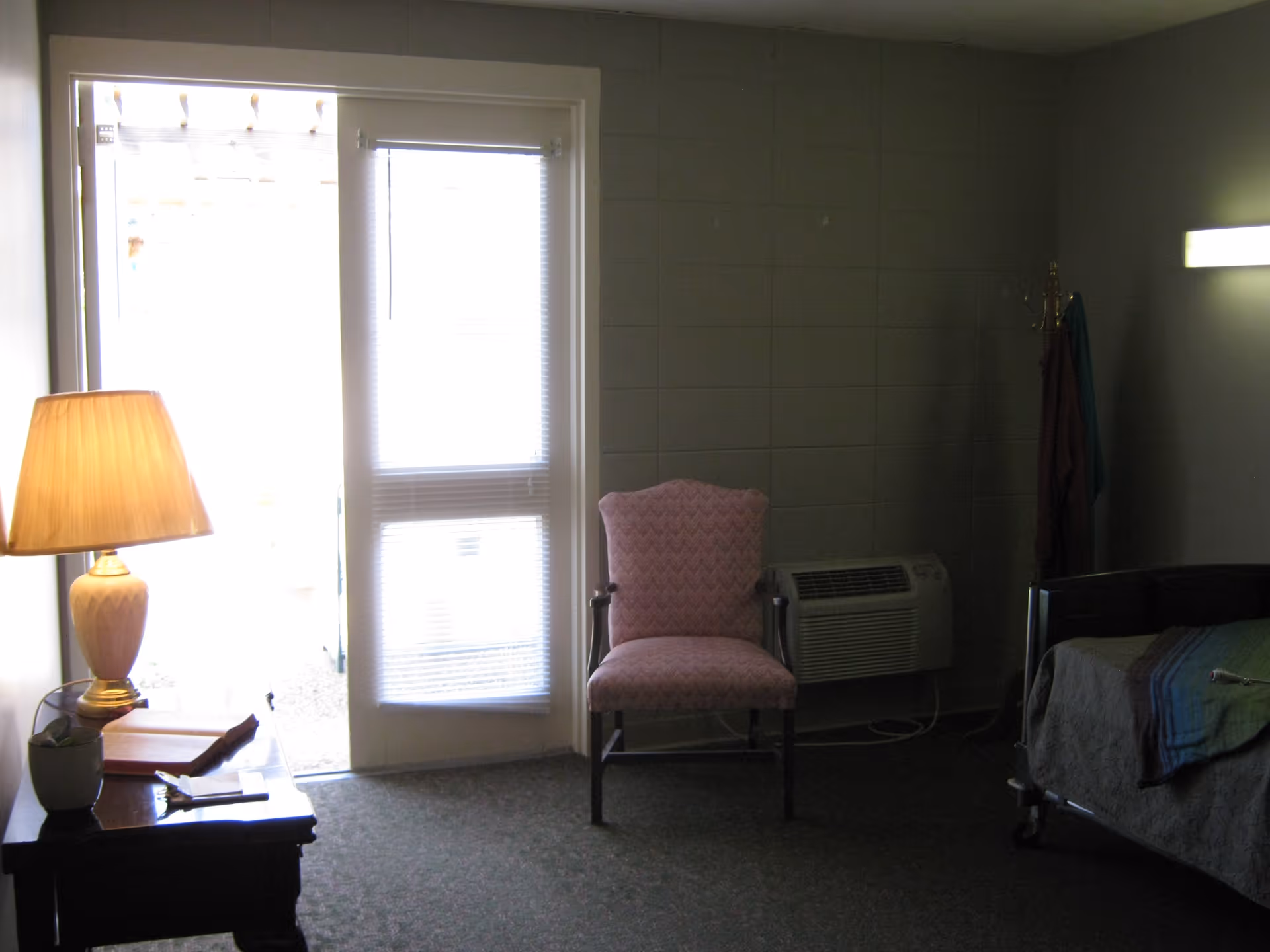 A dimly lit senior living bedroom with a lamp on a bedside table, a pink upholstered chair, a bed, and a glass door with blinds.