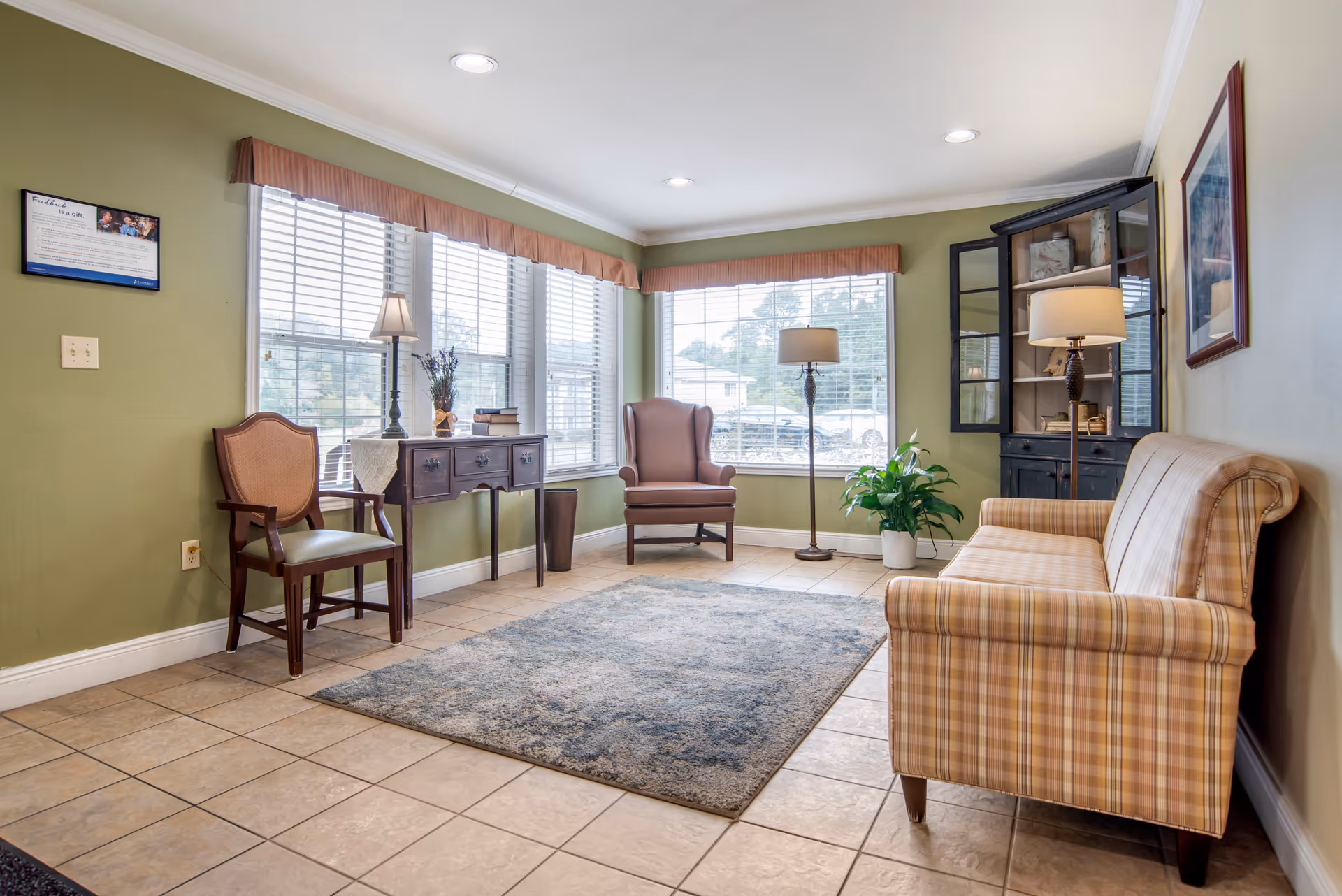 Sunny sitting room with a plaid sofa, armchairs, side table, lamps and a rug in front of large windows.