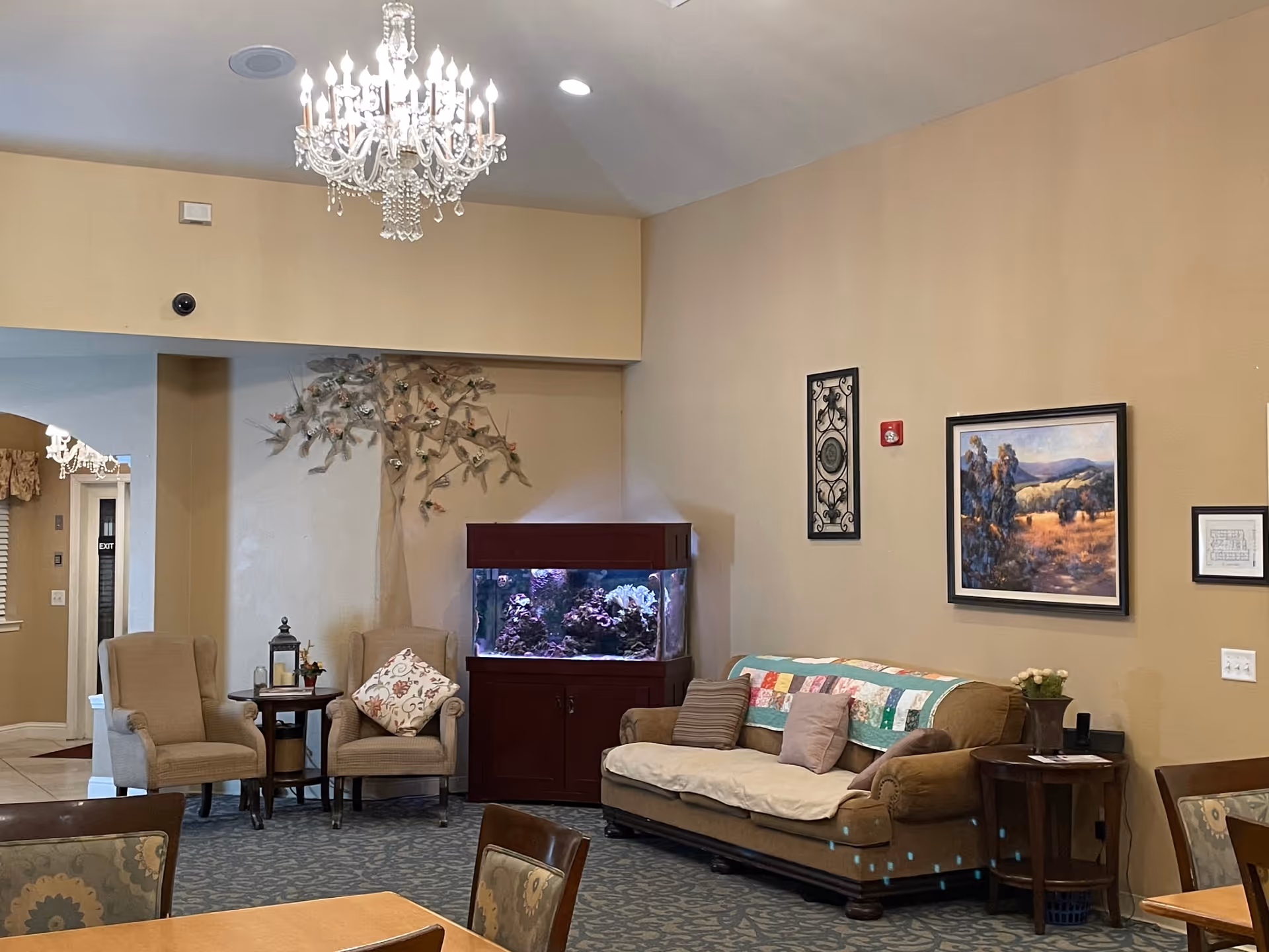 A cozy senior living facility common area with a chandelier hanging from the ceiling, two beige armchairs with a small round table between them, a large fish tank on a wooden stand, and a brown sofa with cushions and a colorful quilt draped over the back. The walls are decorated with framed artwork and a decorative metal wall hanging.