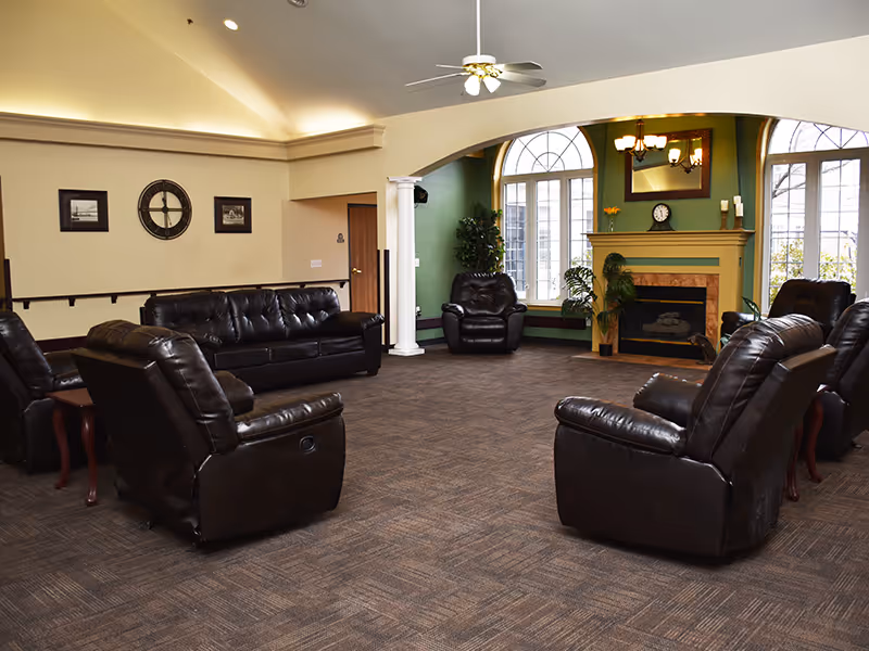 Spacious assisted living common room with leather sofas and armchairs arranged around a fireplace and large arched windows.