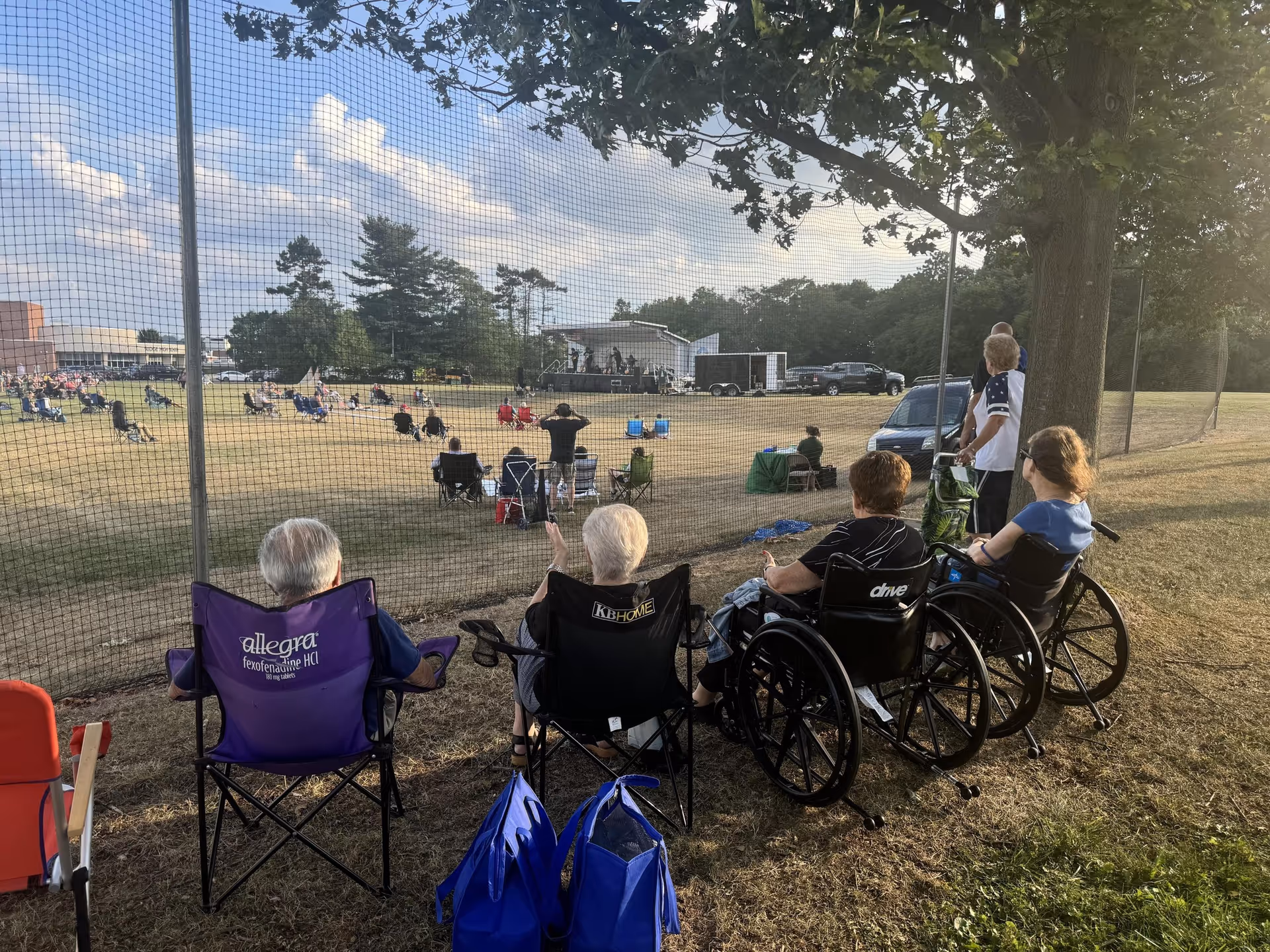 Residents seated in lawn chairs and wheelchairs watch an outdoor concert across a grassy field.