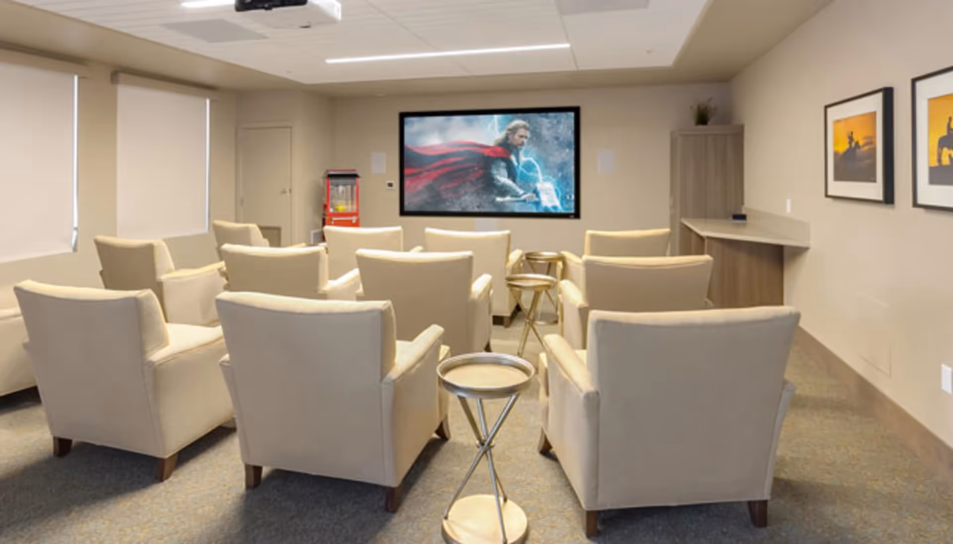 A small theater room with beige armchairs arranged in rows facing a large screen displaying a movie scene featuring a character with a red cape and lightning. The room has beige walls, a popcorn machine in the corner, a projector mounted on the ceiling, and framed artwork on the right wall.