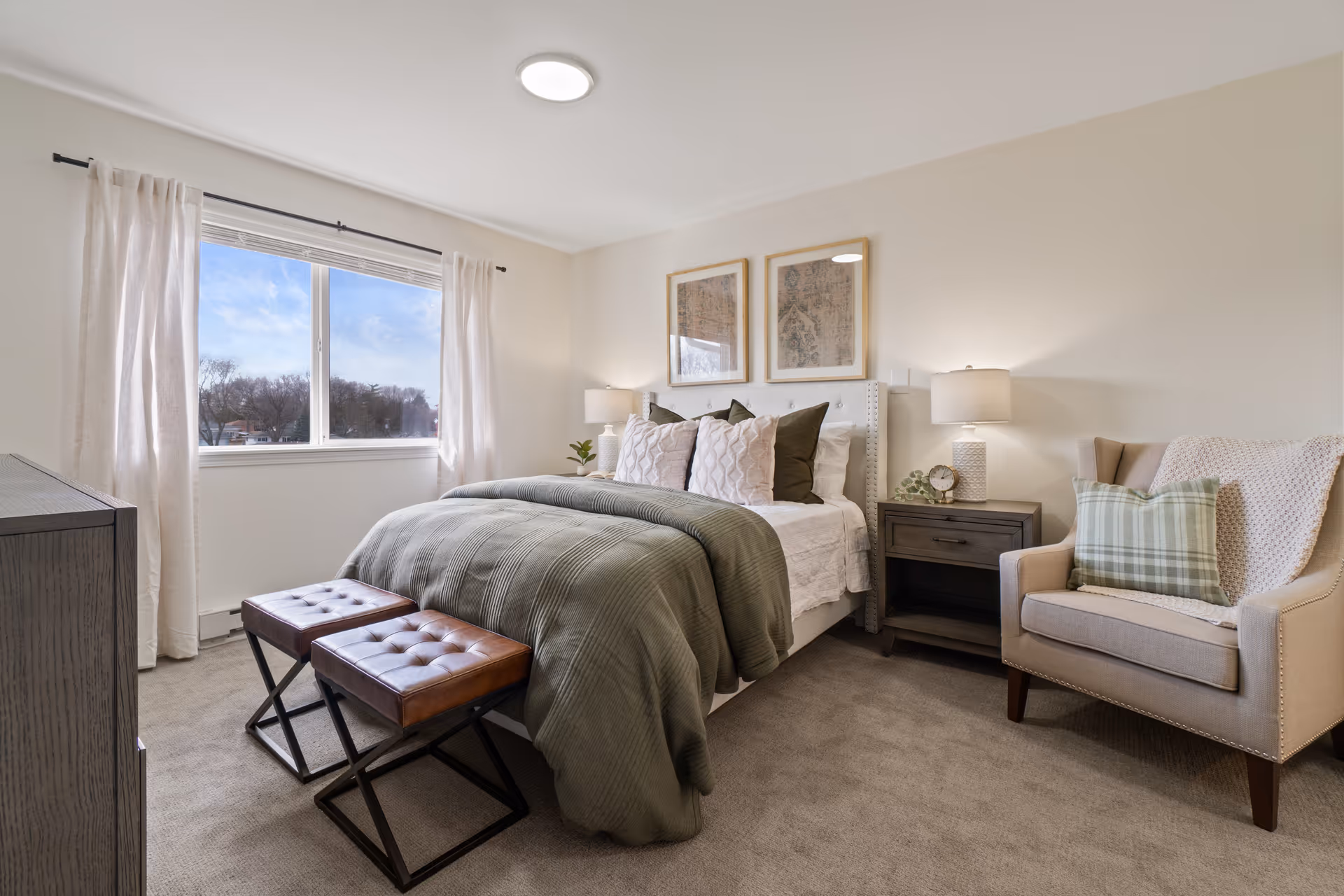 A cozy bedroom with a large window letting in natural light. The bed is made with white sheets, multiple pillows, and a greenish-gray blanket. Two brown leather stools with black metal legs are placed at the foot of the bed. There is a wooden nightstand with a lamp, a small clock, and a plant. A beige armchair with a green plaid pillow and a knitted throw is positioned next to the nightstand. Two framed artworks hang above the bed on the light-colored walls.
